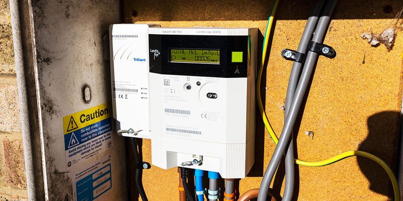 A Landis+Gyr smart electricity meter with a Trilliant communications hub mounted on an outside wall next to a caution label and cabling