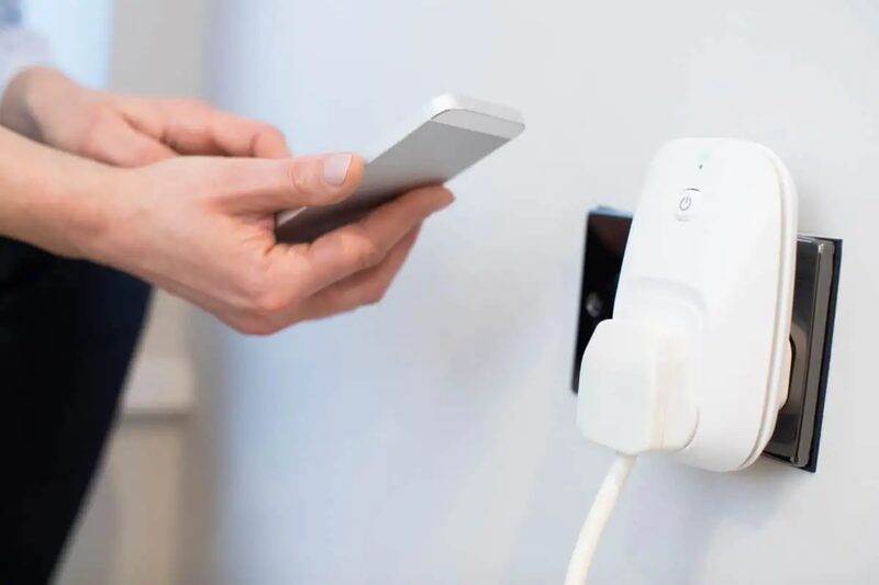 A woman using a smartphone app to control a white smart plug plugged into a UK wall socket