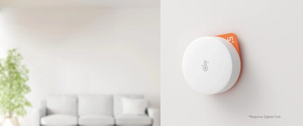 A white SONOFF SNZB-02P Zigbee temperature and humidity sensor mounted on a wall beside a blurred living room