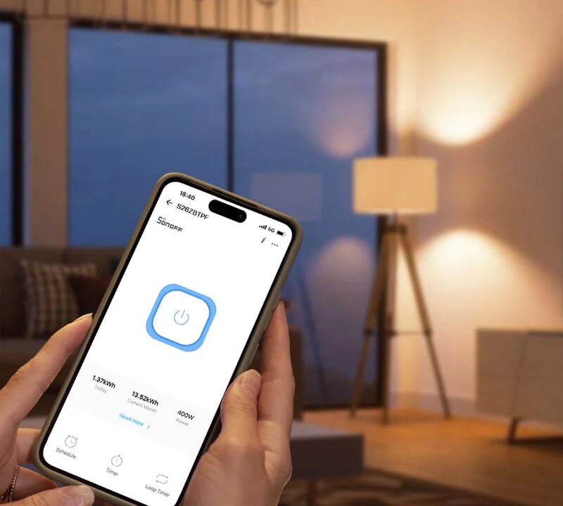 A hand holding a smartphone showing the Sonoff app controlling a smart plug, in a dimly lit living room