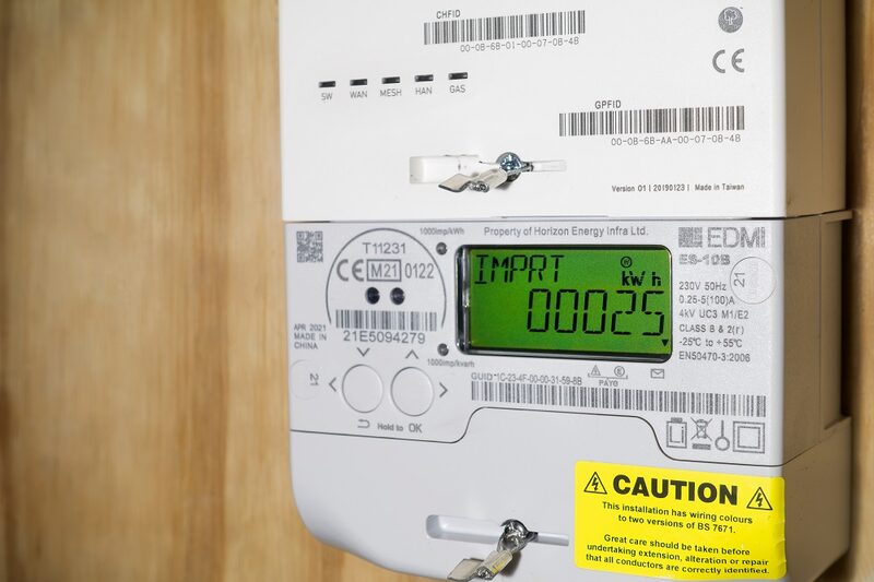 A close-up of an EDMI smart electricity meter with a green LCD display and a communications hub mounted above it on a wooden wall