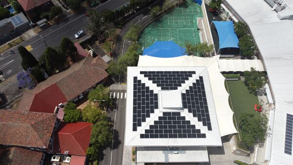 Aerial view of a large rooftop solar panel installation on a building with a pyramid-shaped roof, surrounded by residential houses and a sports court