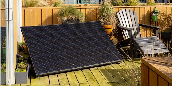 A plug-in solar panel propped at an angle on a wooden balcony terrace next to an Adirondack chair and potted plants