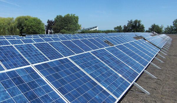 Rows of solar panels mounted on angled metal frames on a flat roof, with trees and blue sky behind