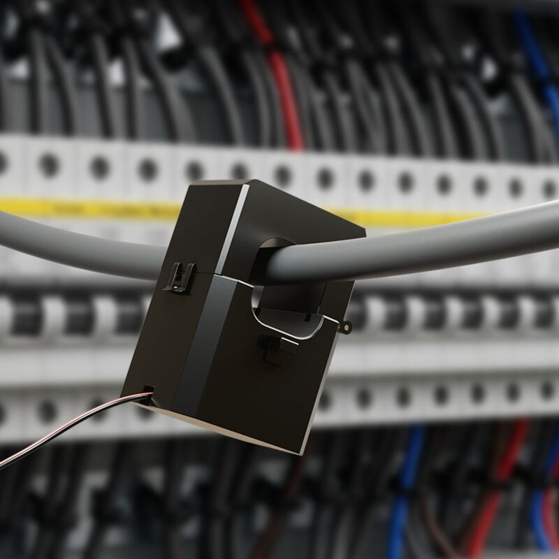 A black CT clamp current transformer clipped around a grey electrical cable in a consumer unit, with a thin signal wire leading out