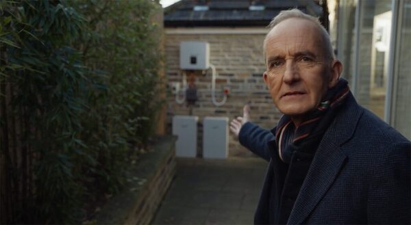 A man gesturing towards a Fox ESS battery and inverter installation on a brick wall outside a house