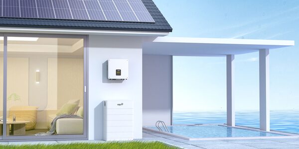 A Fox ESS hybrid inverter and battery mounted on the wall of a modern house with rooftop solar panels, shown in a rendered seaside setting
