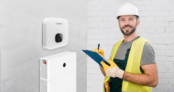 A smiling installer in a hard hat and hi-vis vest with a clipboard next to a wall-mounted Growatt battery inverter and stacked battery modules