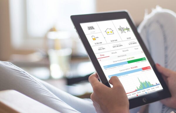 A person holding a tablet displaying a solar PV monitoring app with energy production and consumption charts