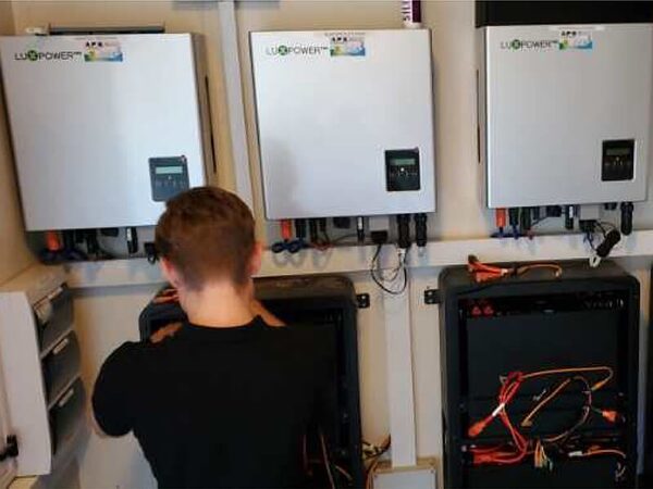 A technician working on wiring below three wall-mounted LuxPower hybrid solar inverters with exposed battery units and cables