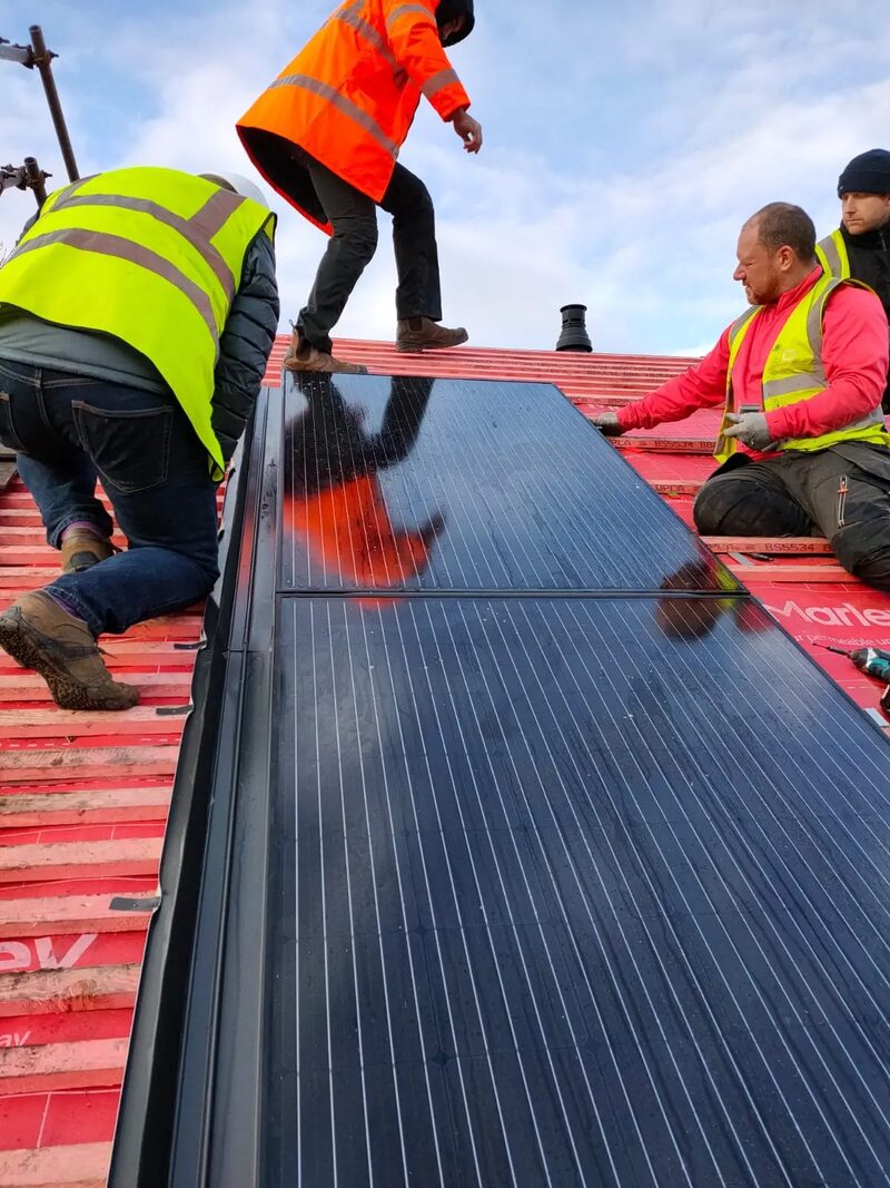 Workers in high-visibility vests installing black Marley solar PV roof tiles on a red-battened roof