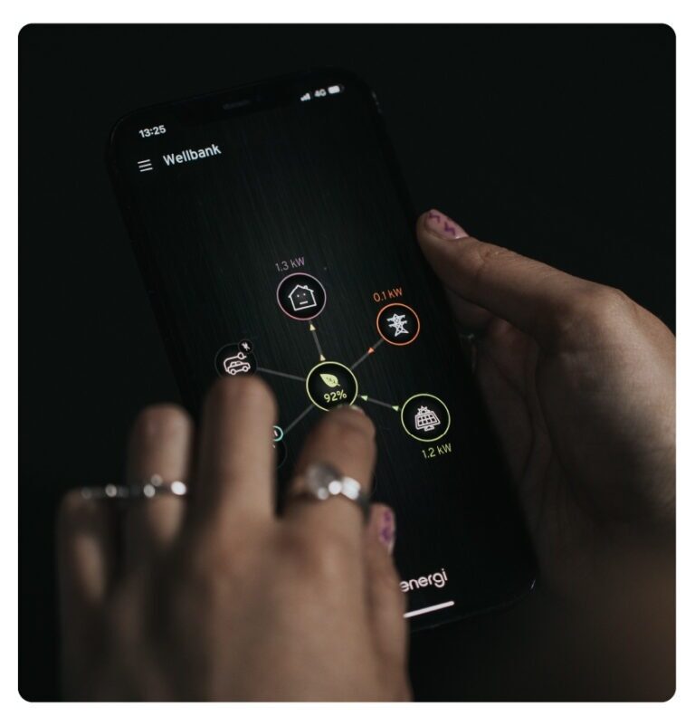 A person's hands holding a smartphone displaying the myenergi app energy monitoring screen with power flow icons