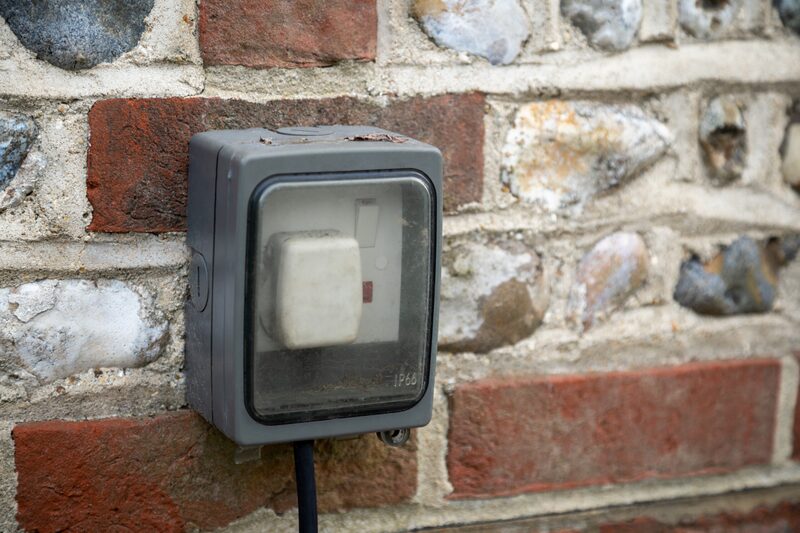 A weatherproof outdoor electrical socket box with a plug and cable mounted on a brick and flint wall