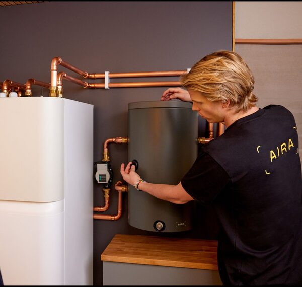 A technician in an Aira t-shirt installing a wall-mounted Aira hot water cylinder connected by copper pipework next to a white heat pump indoor unit