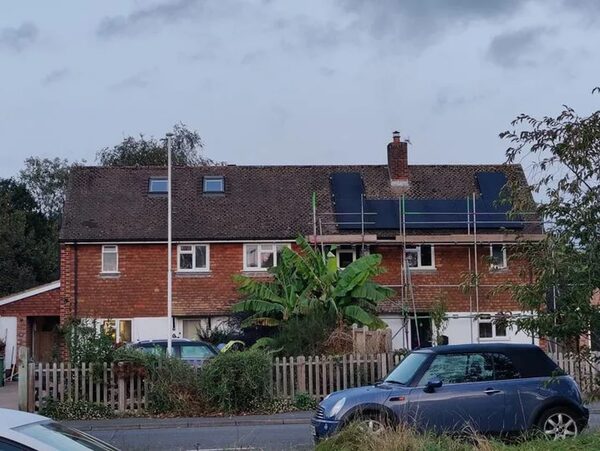 A brick house with solar panels being installed on its tiled roof, surrounded by scaffolding, with cars parked on the street in front.
