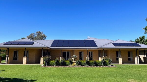 A large single-storey brick house with solar panels installed across its grey metal roof under a clear blue sky