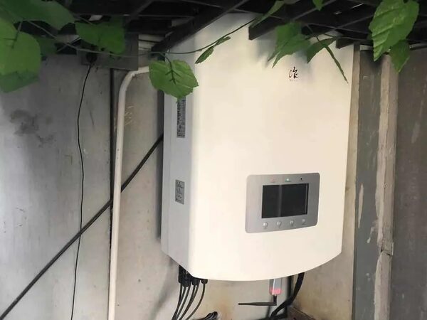A white wall-mounted solar hybrid inverter with a display screen installed on a rough concrete wall with cabling and artificial ivy above it