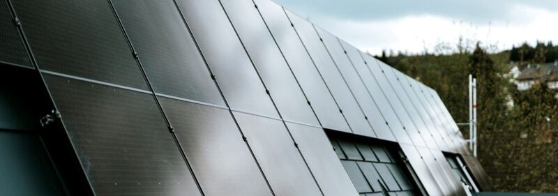 A close-up of dark solar panels mounted on a roof with trees and houses in the background