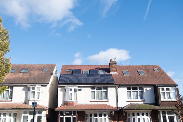 A row of British semi-detached houses under a blue sky, with one roof fitted with solar PV panels.