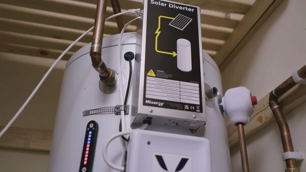 A Mixergy solar diverter unit mounted on a white hot water cylinder with copper pipework in a loft or airing cupboard