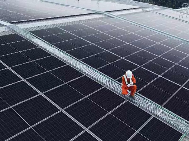 A worker in orange overalls and a white hard hat inspecting a large rooftop solar panel array