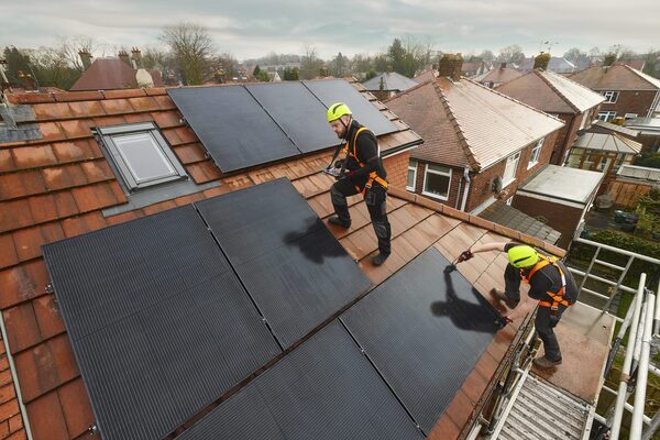 Two workers in safety harnesses and helmets installing solar panels on the tiled roof of a UK house