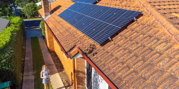 Solar panels installed on the terracotta-tiled roof of a brick house, with a woman standing in the driveway below