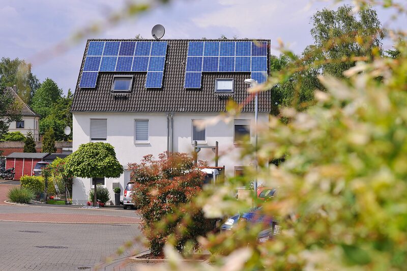 A white semi-detached house with solar panels on its tiled roof, seen from a leafy street