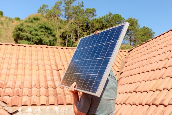 A worker carrying a large solar panel across a terracotta-tiled roof