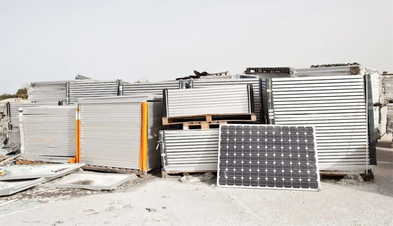 A pile of old solar panels stacked on pallets at an outdoor recycling site, with one panel leaning upright in front