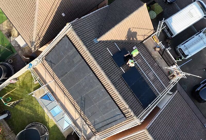 Aerial view of installers in hi-vis fitting black solar panels on the tiled roof of a UK house surrounded by scaffolding