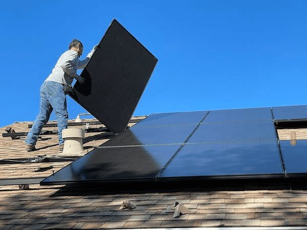 A worker lifting a solar panel onto a shingled roof where several panels are already installed