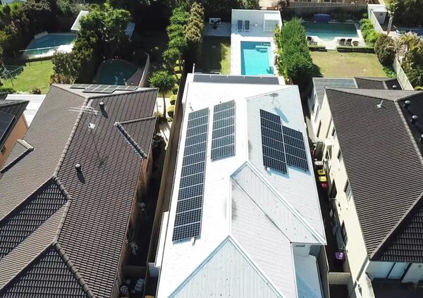 Aerial view of a white suburban house roof covered with solar panels, surrounded by neighbouring roofs and swimming pools