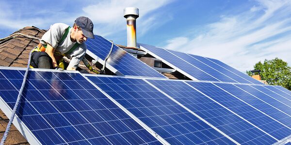A man in a cap and safety harness installing solar panels on a shingled roof next to a chimney flue