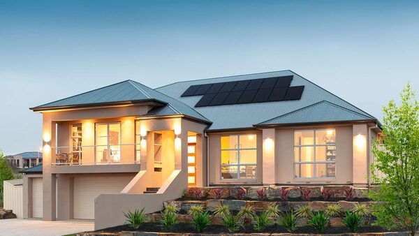 A modern two-storey house with solar panels on its metal roof at dusk, with illuminated windows and landscaped garden
