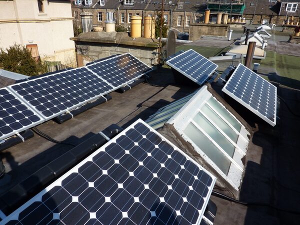 Several solar panels mounted on a flat urban rooftop with skylights and tenement buildings behind