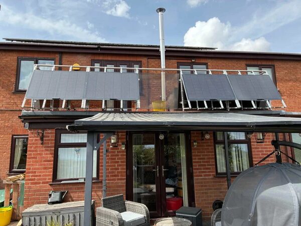 Solar panels mounted on a rack above a flat roof extension of a brick house, seen from the garden