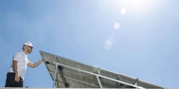 An installer in a white hard hat inspecting a solar panel array against a bright blue sky