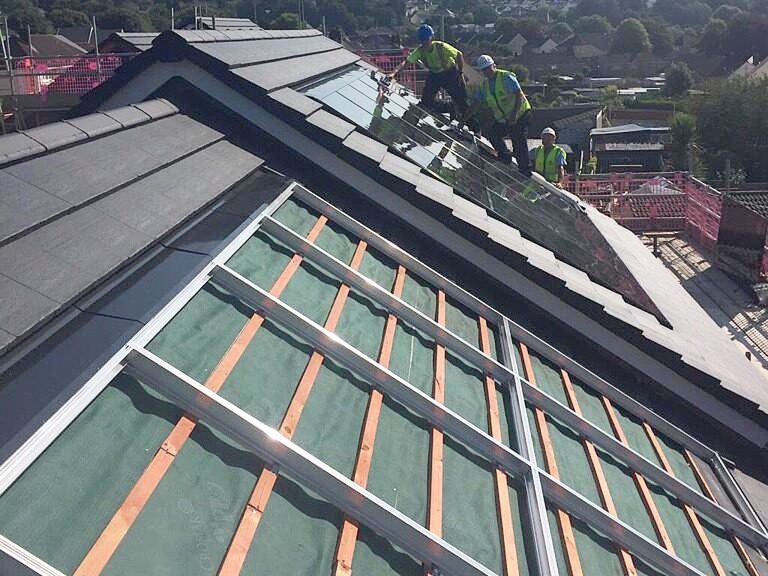 Roofers installing integrated solar panels on a tiled house roof, with mounting rails and battens visible on the unfinished section