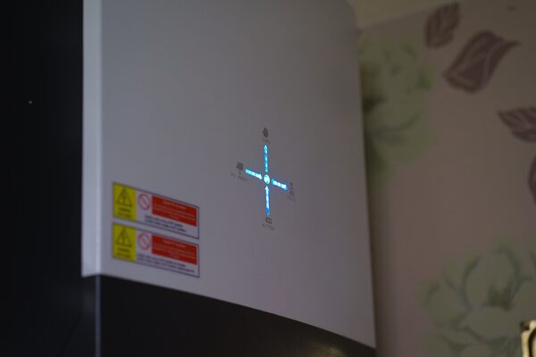 A white solar inverter mounted on a wall indoors, with blue indicator lights in a plus shape and warning labels
