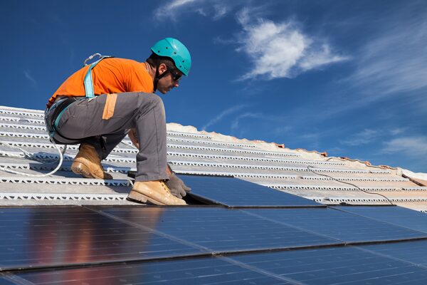 A worker in a hard hat and harness installing solar panels on a tiled roof under a blue sky