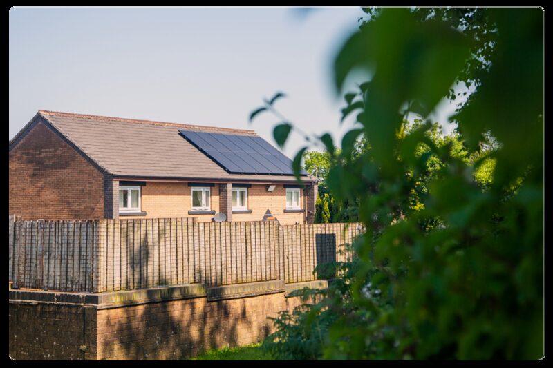 A brick semi-detached house with solar panels on its roof, seen behind a wooden fence and greenery