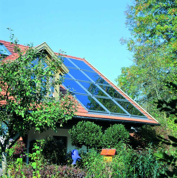 A house with dark solar thermal collector panels fitted on its red tiled roof, surrounded by trees and garden