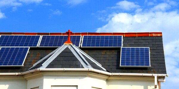 Solar panels mounted on the slate roof of a house with a bay window, against a blue sky