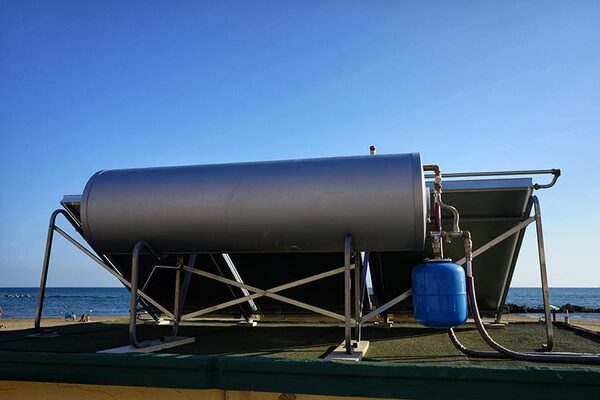 A rooftop thermosyphon solar water heater with a horizontal cylinder tank and flat-plate collector panels, photographed by the sea
