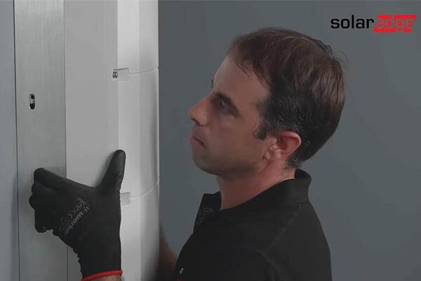 A technician in black gloves mounting a white SolarEdge Home Hub inverter on a wall