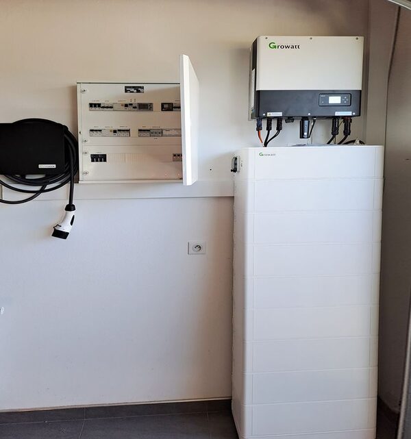 A Growatt hybrid inverter mounted on a wall above a tall white Growatt home battery, next to an open consumer unit and an EV charging cable