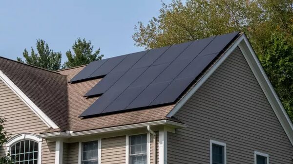 A house with black solar panels installed on its shingled roof under a blue sky with trees behind