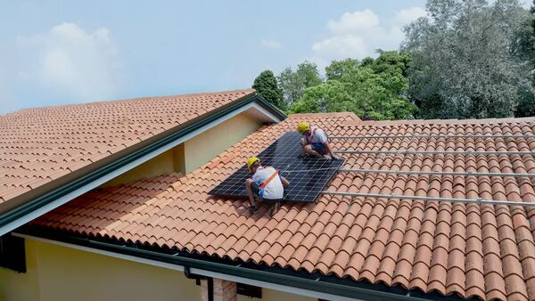 Two workers in safety harnesses and helmets installing a solar panel on a terracotta tiled roof
