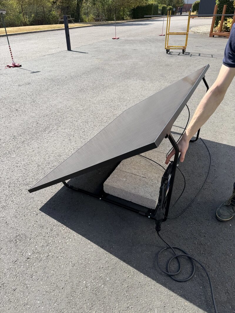 A person handling a ground-mounted solar panel on a black metal frame with a cable, set up on a tarmac yard